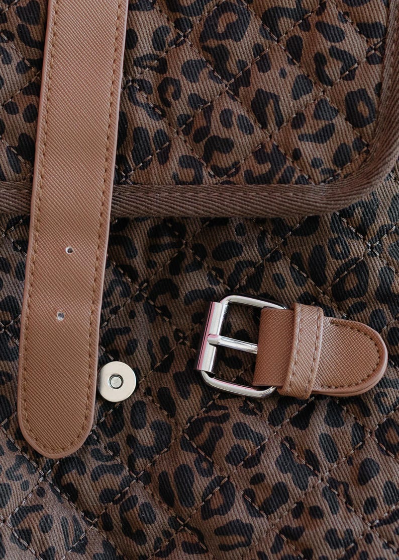 K&ouml;nnte beinhalten: Nahaufnahme eines braun-schwarzen Stoffes mit Leopardenmuster und Steppmuster. Ein hellbrauner Riemen mit silberner Schnalle und Magnetverschluss ist sichtbar. Der Stoff ist dunkelbraun mit schwarzen Flecken.