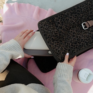 K&ouml;nnte beinhalten: Ein wei&szlig;es und graues elektronisches Ger&auml;t wird neben einer schwarzen Leopardenmuster-H&uuml;lle mit einer braunen Schnalle gehalten. Ein rosa Pullover und ein wei&szlig;er und rosa Becher sind ebenfalls sichtbar. Die Artikel befinden sich auf einer hellen Oberfl&auml;che.