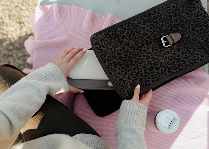 K&ouml;nnte beinhalten: Ein wei&szlig;es und graues elektronisches Ger&auml;t wird neben einer schwarzen Leopardenmuster-H&uuml;lle mit einer braunen Schnalle gehalten. Ein rosa Pullover und ein wei&szlig;er und rosa Becher sind ebenfalls sichtbar. Die Artikel befinden sich auf einer hellen Oberfl&auml;che.