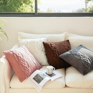 Peut inclure: Quatre coussins de canapé en tricot de différentes couleurs et motifs sur un canapé blanc. Les coussins sont rose, blanc, marron et gris. Une tasse en céramique blanche et un livre sont sur le canapé.