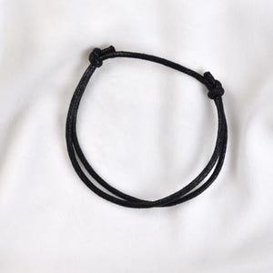 May include: A simple black bracelet made of two cords, knotted at the ends. The bracelet is displayed on a white, textured surface. This minimalist accessory is suitable for everyday wear.