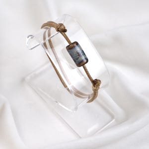 May include: A handcrafted bracelet with a cylindrical bead, patterned in grey and brown. It's strung on a tan cord, fastened with knots. The bracelet is displayed on a clear acrylic stand against a white backdrop.