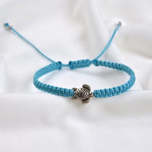 May include: A turquoise macrame bracelet with a silver turtle charm. The bracelet is adjustable with sliding knots and is displayed on a white, textured background. The turtle charm is detailed and adds a nautical theme.