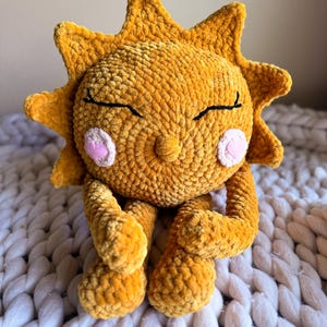 Puede incluir: Un juguete de peluche de sol tejido a crochet con textura de terciopelo amarillo. El sol tiene una cara sonriente con los ojos cerrados y mejillas rosadas.