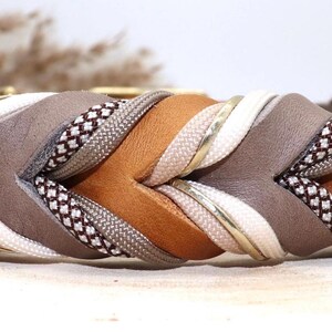 May include: A braided dog collar with a brown, gray, white, and gold color scheme. The collar is made of leather and fabric and has a woven design.