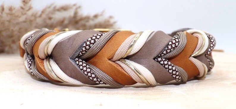 May include: A braided dog collar with a brown, gray, white, and gold color scheme. The collar is made of leather and fabric and has a woven design.