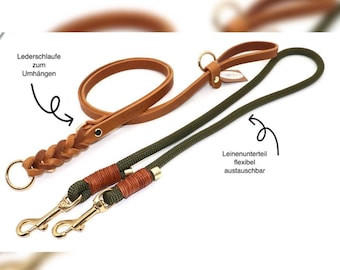May include: A brown leather dog leash with a braided leather handle and a green rope leash with two gold metal clips. The leather leash has a loop for hanging. The text "Lederschlaufe zum Umhängen" is written on the image. The text "Leinenunterteil flexibel austauschbar" is written on the image.