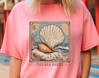 Puede incluir: Camiseta rosa con un gráfico de una concha en una ilustración de estilo vintage. El gráfico está rodeado por un marco marrón y el texto "THE SEA SHELL" está debajo de la imagen.