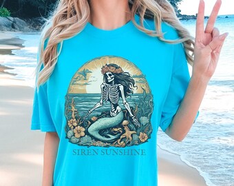 May include: A turquoise blue t-shirt with a graphic of a skeleton mermaid with a flower crown sitting on a rock in the ocean. The text "SIREN SUNSHINE" is printed below the graphic.