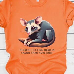 May include: Orange t-shirt with a cartoon opossum and the text "Because playing dead is easier than adulting."