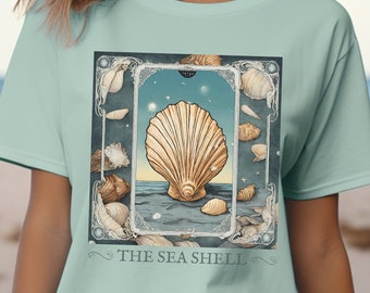 Puede incluir: Una camiseta azul claro con un estampado gráfico de una concha en un marco decorativo. El texto "THE SEA SHELL" está impreso debajo de la imagen.