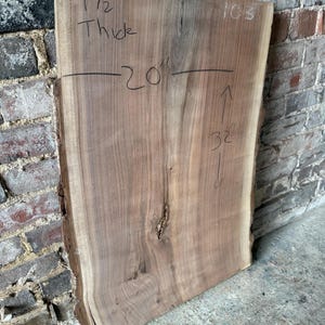 Könnte beinhalten: Eine rohe, unfertige Holzplatte mit natürlichen Kanten. Die Platte ist etwa 3,8 cm dick, 50,8 cm breit und 81,3 cm lang. Das Holz hat eine warme braune Farbe mit sichtbarer Maserung und einem Ast. Die Nummer "103" ist rechts oben geschrieben.