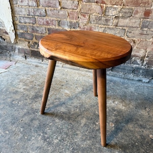 Może przedstawiać: Mały drewniany stołek z okrągłym, owalnym blatem i trzema nogami. Stołek jest wykonany z jasnobrązowego drewna i ma naturalne wykończenie.
