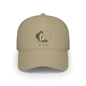 Può includere: Un cappello da baseball color cachi con un'immagine ricamata di un pesce persico verde che salta fuori dall'acqua con il testo "Kiss my Bass" sotto.