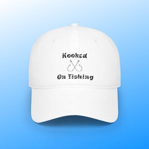 Può includere: Cappellino da baseball bianco con il testo "hooked on fishing" e un'immagine di due ami da pesca incrociati.