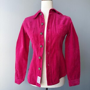 Puede incluir: Una camisa de pana fucsia vibrante con mangas largas, cuello y cierre de botones. La camisa tiene dos bolsillos en el pecho y se muestra en un maniquí. La tela tiene un aspecto texturizado y acanalado.