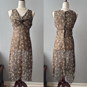 May include: A sheer brown dress with a floral pattern, displayed on a mannequin. The dress features a V-neckline with a bow detail and a flowing, asymmetrical hemline. The dress is sleeveless and has a button closure in the back.