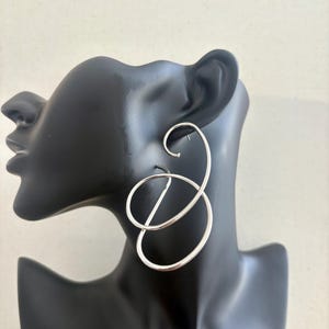 May include: Silver-coloured, abstract hoop earrings. The earrings have a looping design with a curved top and a circular element, displayed on a black mannequin head. The earrings have a polished finish and a modern aesthetic.