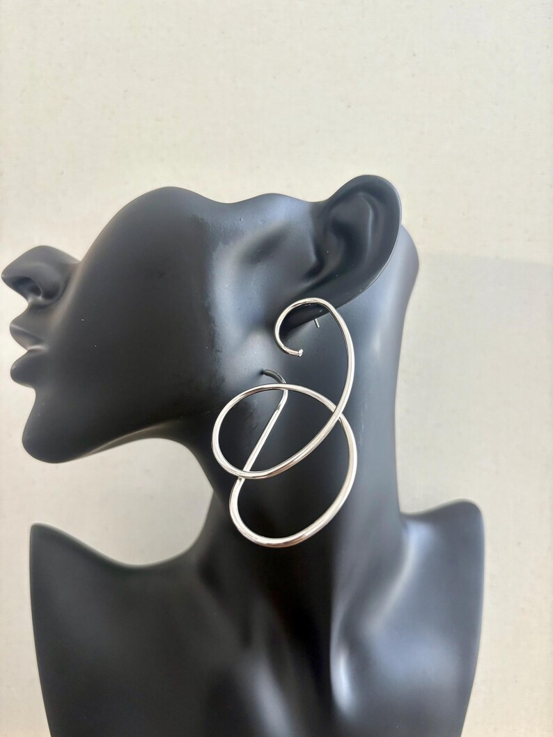 May include: Silver-coloured, abstract hoop earrings. The earrings have a looping design with a curved top and a circular element, displayed on a black mannequin head. The earrings have a polished finish and a modern aesthetic.