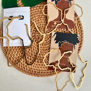 May include: Gold-toned metal earrings in the shape of the African continent. The earrings are displayed on a woven basket with a white background. The earrings are from Manera Collections.