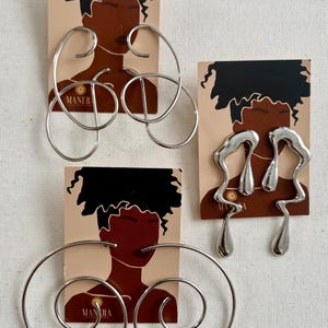 May include: Three pairs of silver earrings are shown against a beige backdrop. The earrings have distinctive designs, including abstract shapes and spiral forms. Each pair is displayed on a card with a graphic of a person's face and the word "MANERA".