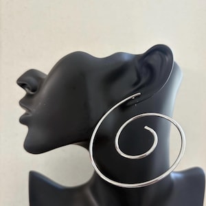 May include: A pair of large, silver spiral hoop earrings. The earrings are displayed on a black mannequin head against a neutral background. The spiral design creates a bold, modern look, suitable for everyday wear or special occasions.
