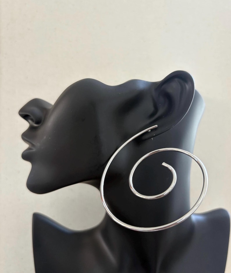 May include: A pair of large, silver spiral hoop earrings. The earrings are displayed on a black mannequin head against a neutral background. The spiral design creates a bold, modern look, suitable for everyday wear or special occasions.