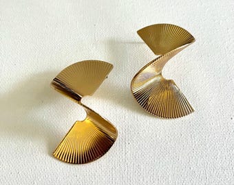 May include: Gold-toned, abstract earrings with a fan-like design. The earrings have a curved, spiral shape with a textured, ridged surface. The earrings are set against a white background.