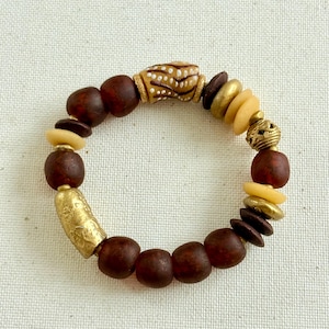 Puede incluir: Un brazalete de cuentas con una variedad de cuentas en tonos marrón, dorado y crema. El brazalete incluye cuentas redondas, cilíndricas y en forma de disco, creando una textura y un diseño únicos. El brazalete se muestra sobre un fondo neutro.
