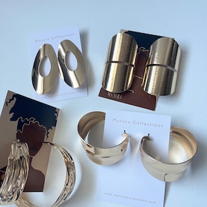 May include: A collection of gold-toned earrings, including large oval drop earrings, wide cuff earrings, and thick hoop earrings. The earrings are displayed on white cards with the brand name "Manera Collections" and website address.