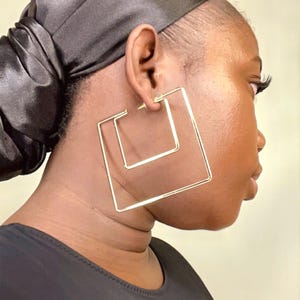 May include: Gold-toned square hoop earrings. The earrings are a double-layered square design. The image shows a close-up of the earrings on a person's ear. The person is wearing a black head covering and a black top.