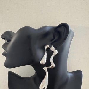 May include: A pair of silver earrings with an abstract, flowing design. The earrings have a polished, reflective surface and are displayed on a black mannequin head. The design is organic and modern, with a unique, sculptural shape, set against a neutral backdrop.