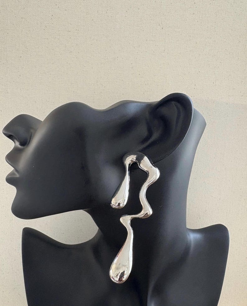 May include: A pair of silver earrings with an abstract, flowing design. The earrings have a polished, reflective surface and are displayed on a black mannequin head. The design is organic and modern, with a unique, sculptural shape, set against a neutral backdrop.