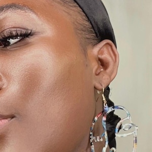 May include: Large, abstract face-shaped earrings with a colorful, marbled pattern. The earrings are made of a translucent material and feature a cutout design with gold-colored hooks. The earrings are a unique fashion accessory.