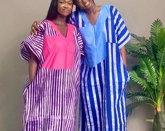 May include: Two women wearing matching V-neck dresses. One dress is pink and purple striped with a pink square on the chest. The other dress is blue and white striped with a blue square on the chest. Both dresses have pockets.