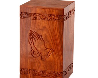 May include: A rectangular wooden urn with a rich, reddish-brown finish. The front features an engraved image of praying hands. Decorative floral patterns adorn the top and bottom edges. The urn is designed for memorial purposes.