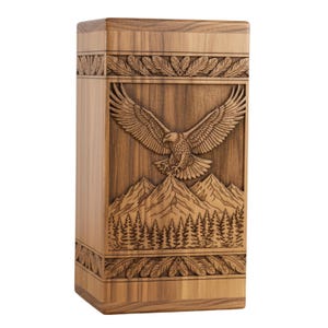 May include: A rectangular wooden urn with a carved design. The design features an eagle with outstretched wings above a mountain range and a forest. The urn is made of light brown wood and has decorative leaf patterns.