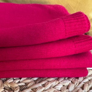 Puede incluir: Primer plano de prendas de punto rosa brillante dobladas. La tela es suave y parece ser un suéter o una prenda similar. El puño acanalado de la manga es visible. El fondo incluye un cojín amarillo y una superficie tejida.