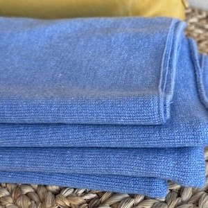 Puede incluir: Una pila de bufandas de punto azul claro dobladas. Las bufandas están cuidadosamente dispuestas, mostrando la suave textura y el color. La imagen resalta las sutiles variaciones de la tela y las líneas limpias de los bordes doblados. El fondo incluye una superficie tejida y un cojín amarillo.
