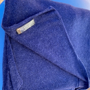 Peut inclure: Une écharpe en cachemire bleu foncé pliée. L'écharpe est faite d'une matière douce et texturée. Une petite étiquette noire est visible, avec le texte "100% Exclusive Cashmere Hand Made in Nepal Dry Clean Only".