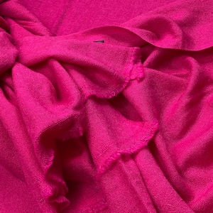 Op de afbeelding: Een close-up van een levendige fuchsia-kleurige stof, mogelijk een sjaal of omslagdoek. Het materiaal is zacht en lijkt geweven, met zichtbare textuur en gerafelde randen. De stof is opgevouwen en geplooid, waardoor schaduwen en diepte ontstaan.