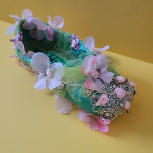 May include: A green satin ballet slipper decorated with pink and white fabric flowers, green tulle, and gold accents.