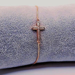 May include: Rose gold bracelet with a cross pendant. The cross is adorned with small, clear gemstones. The bracelet has a fine chain and is shown on a soft, grey surface.