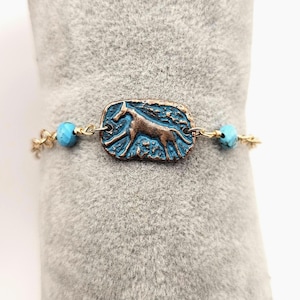 May include: A gold-toned bracelet featuring a rectangular, bronze-colored charm with a horse silhouette against a blue background. The bracelet includes turquoise beads and delicate gold chain links.