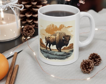 May include: White ceramic mug featuring a detailed illustration of a bison in a natural landscape with mountains and clouds. The mug has a curved handle and is filled with a dark liquid. The design is in shades of brown, beige, and tan.