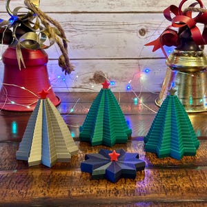 May include: A festive holiday scene featuring three miniature Christmas trees in shades of green and gray, each topped with a red star. Two decorative bells with bows and string lights are in the background. The scene is set on a wooden surface.