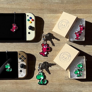 May include: Two Nintendo Switch consoles, one white and one black, with green and red pixelated character charms attached. The consoles are on a wooden surface with small boxes containing additional charms.