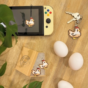May include: A collection of pixelated chicken-themed accessories, including a keychain, earrings, and a charm hanging from a game console. The accessories are displayed with white eggs on a wooden surface. A box with a fox logo is also present.