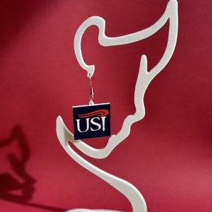 Könnte beinhalten: Ein Paar Ohrringe mit einem quadratischen Anhänger mit den Buchstaben "USI" in Rot und Weiß auf blauem Hintergrund. Der Anhänger hängt an einem silbernen Haken.