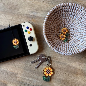 Puede incluir: Una consola Nintendo Switch blanca con un amuleto de girasol sujeto a la pantalla. Un llavero con un amuleto de girasol y un bloque verde se encuentra sobre una superficie de madera. Una cesta de mimbre con dos pendientes de girasol se encuentra en el fondo.
